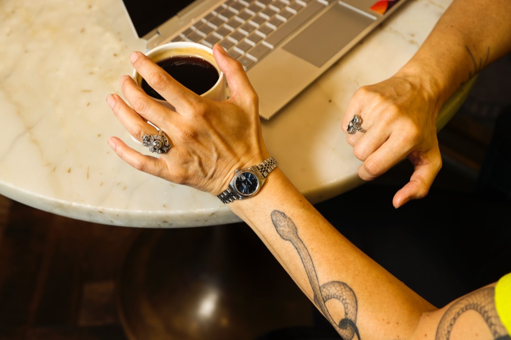 Close-up of Emy Tafelski's hands with silver rings and a snake tattoo, wrapped around a coffee cup next to a laptop