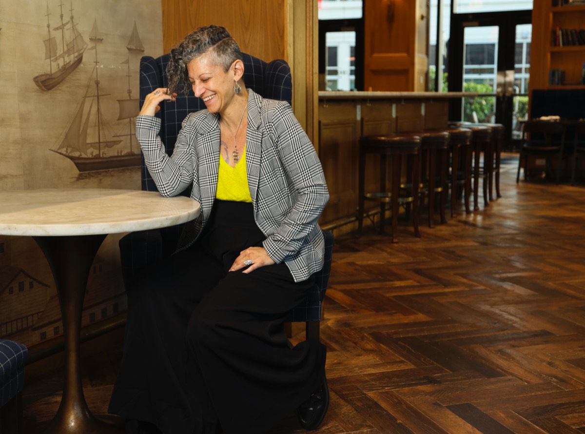 Emy Tafelski smiling down in a houndstooth blazer and yellow top, seated at a marble table in a wood-paneled cafe