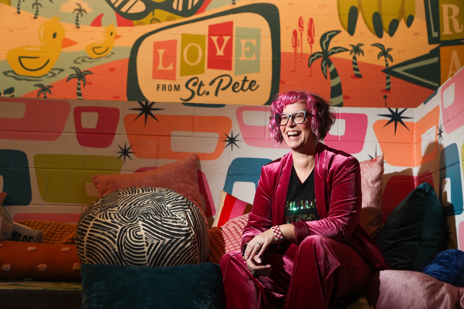 Robyn Sayles laughing in a pink velvet suit on a pile of pillows, under a retro 'Love from St. Pete' mural