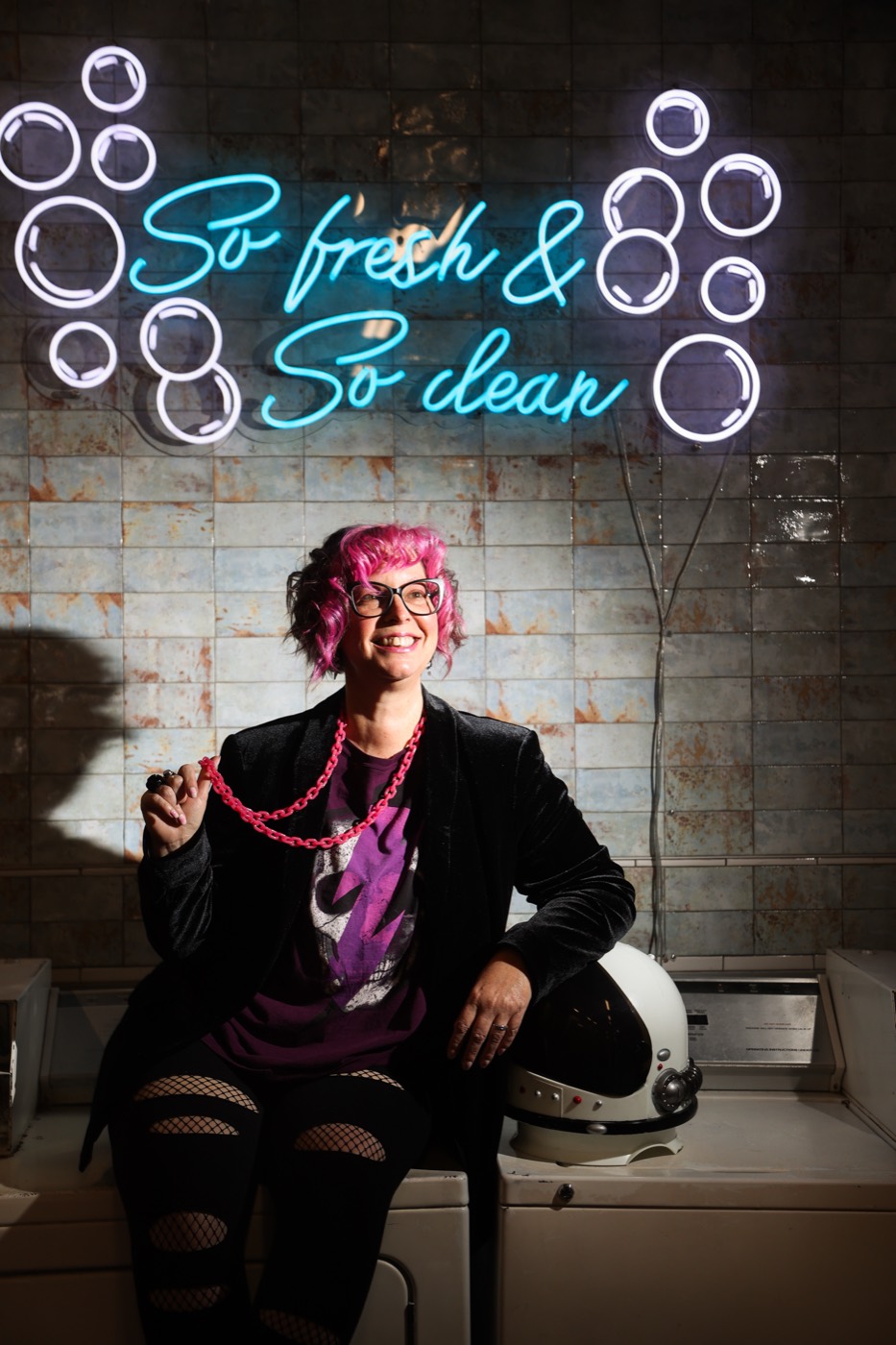 Robyn Sayles sitting on a laundromat washer beneath a neon sign reading 'So fresh and so clean,' holding a pink chain beside a space helmet
