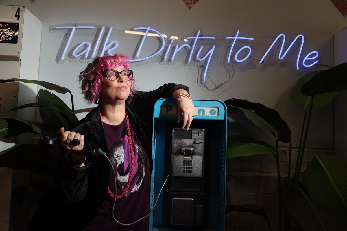 Robyn Sayles leaning on a vintage blue payphone, receiver in hand, under a neon 'Talk Dirty to Me' sign
