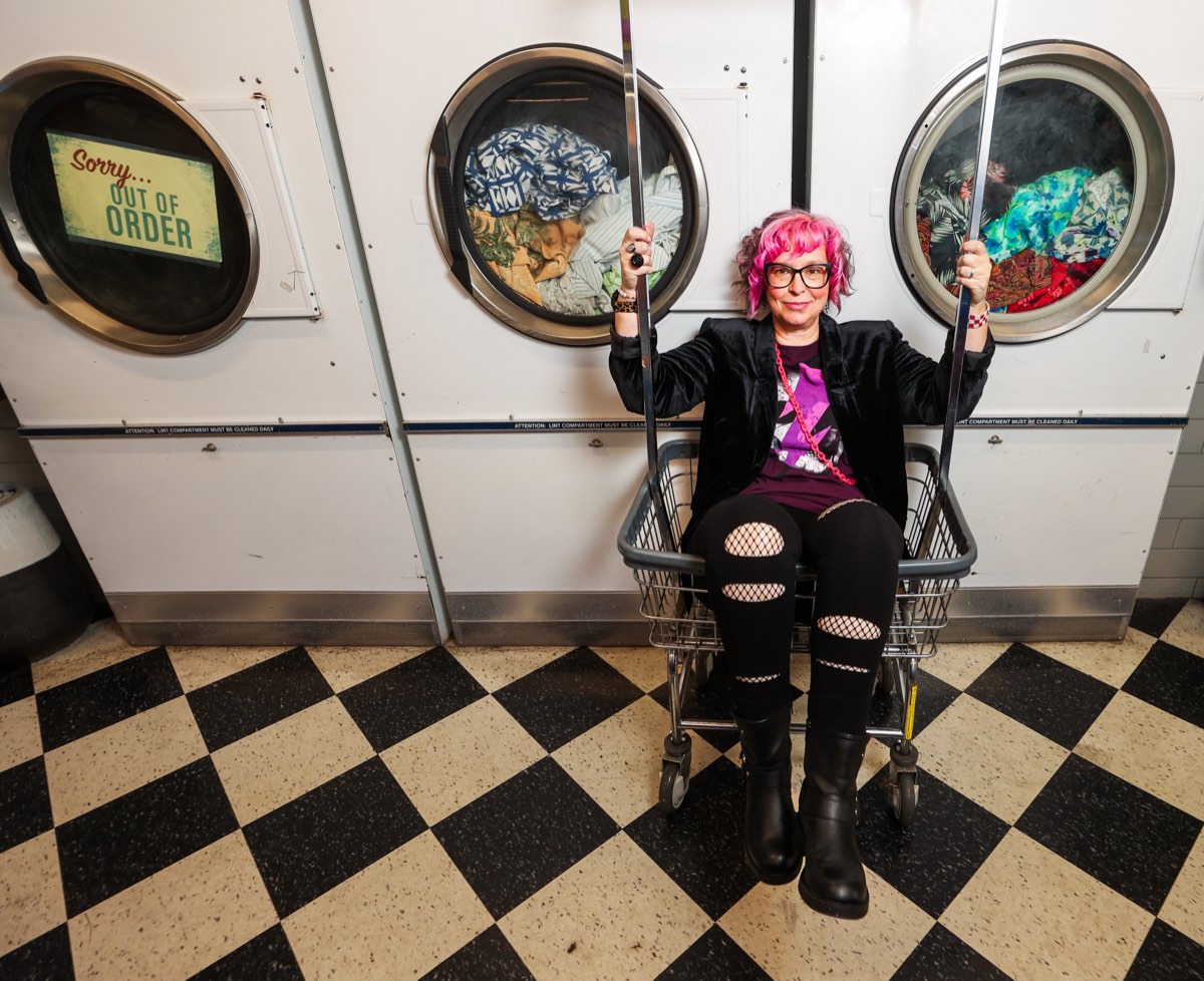 Robyn Sayles riding a laundry cart in front of a row of dryers on a black and white checkered floor