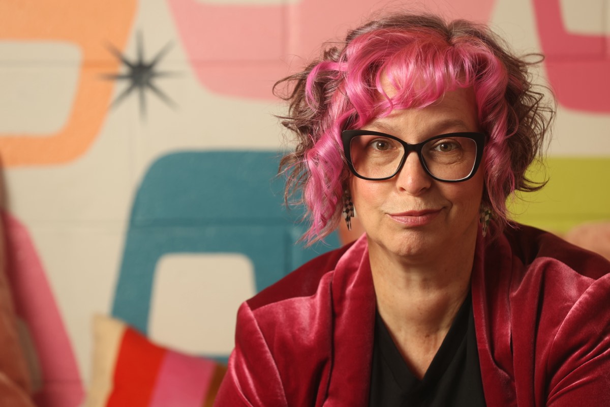 Close-up portrait of Robyn Sayles with pink curly hair and black cat-eye glasses against a colorful mid-century mural